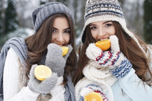two attractive women in winter clothes eating oranges with a winter scene in the background